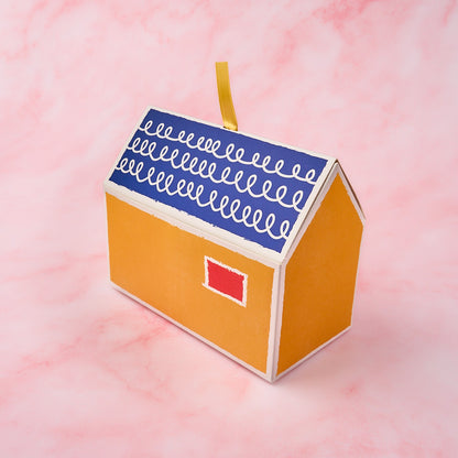 Grah Pravesh return gift box in a yellow house design with blue roof and window detail on a soft pink background