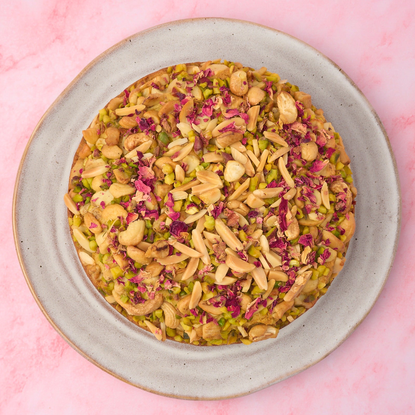 Top view of eggless Pistachio Mawa Cake topped with mixed nuts and dried rose petals by EatWithSimer